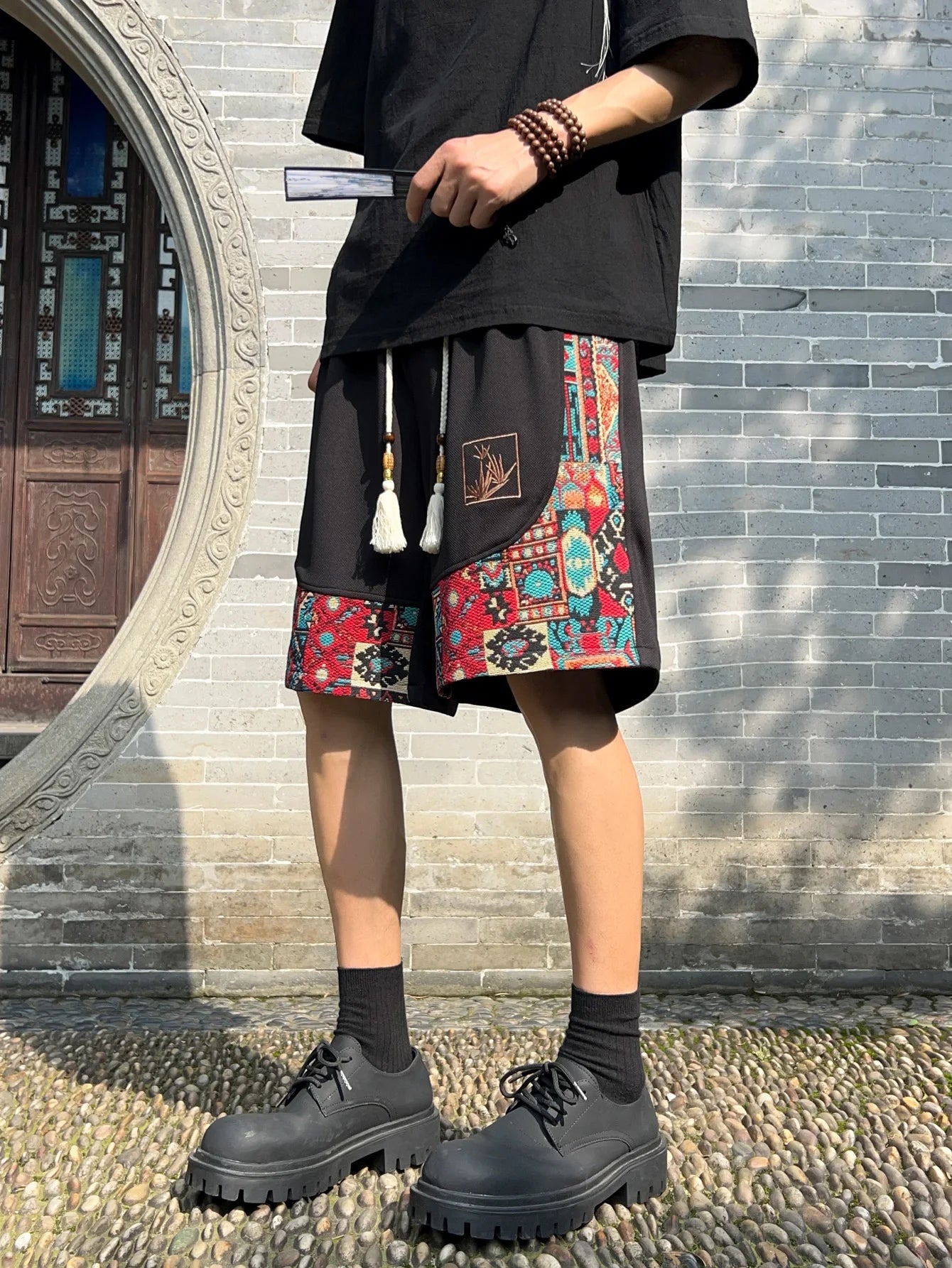 Angled front view of black embroidered patchwork shorts styled with fan and beads
