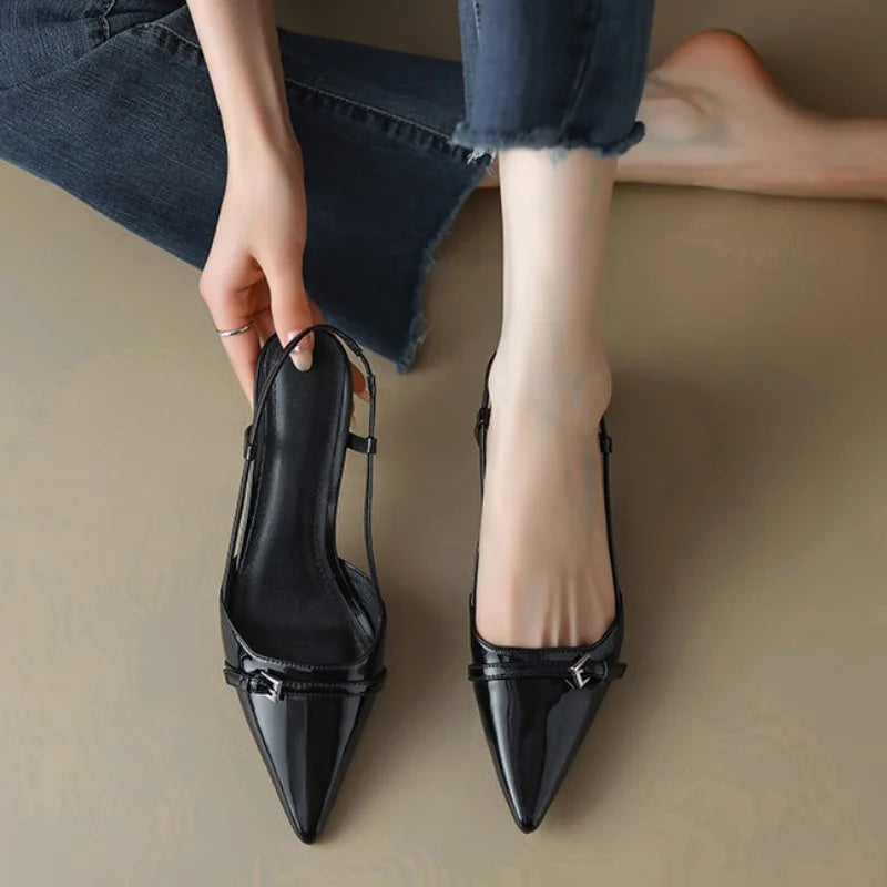 Black patent pointed-toe slingback heels with buckle detail, held in hand