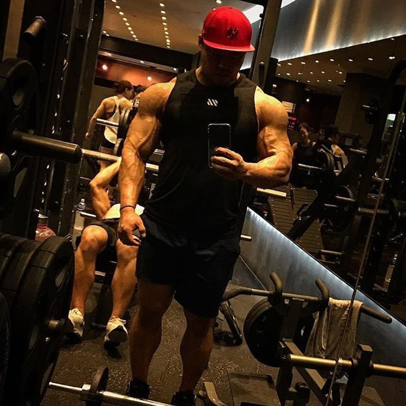 Bodybuilder in black training tank top taking selfie in gym mirror with weights visible