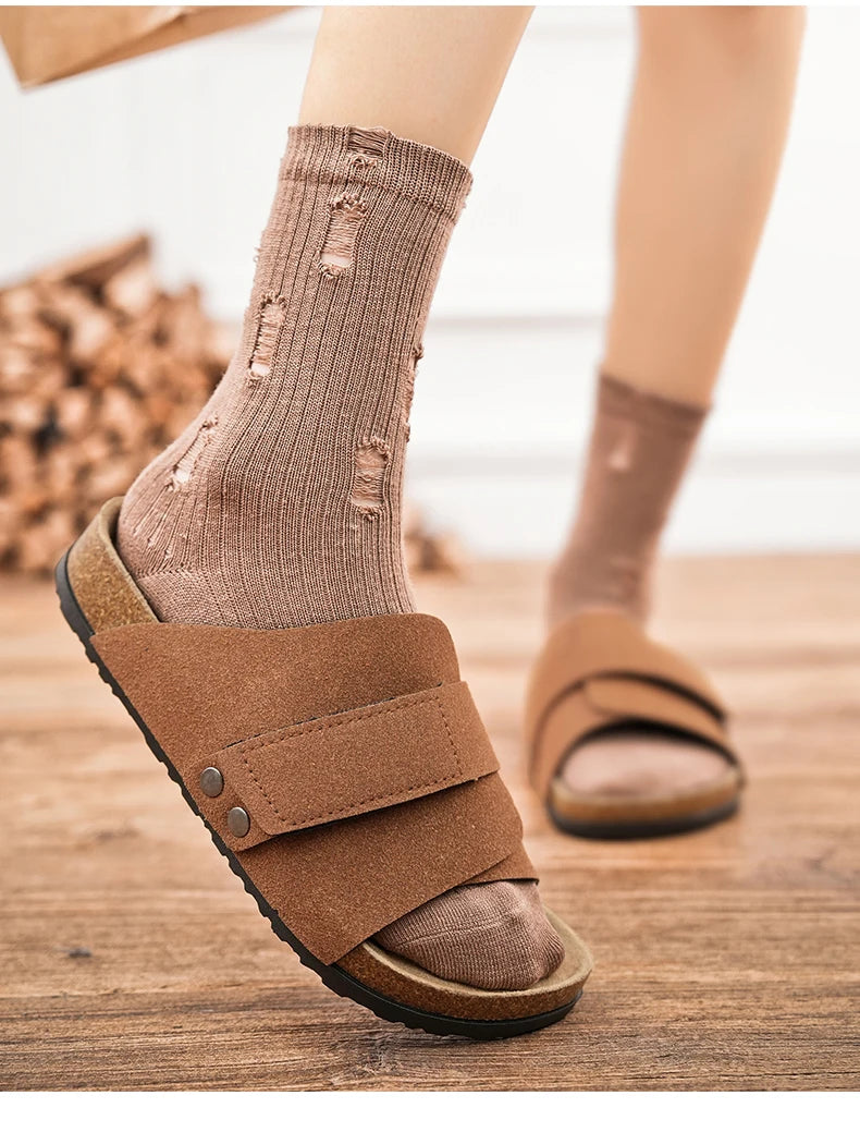 Brown suede sandal worn with distressed brown socks on wooden floor, close-up of footbed and sole