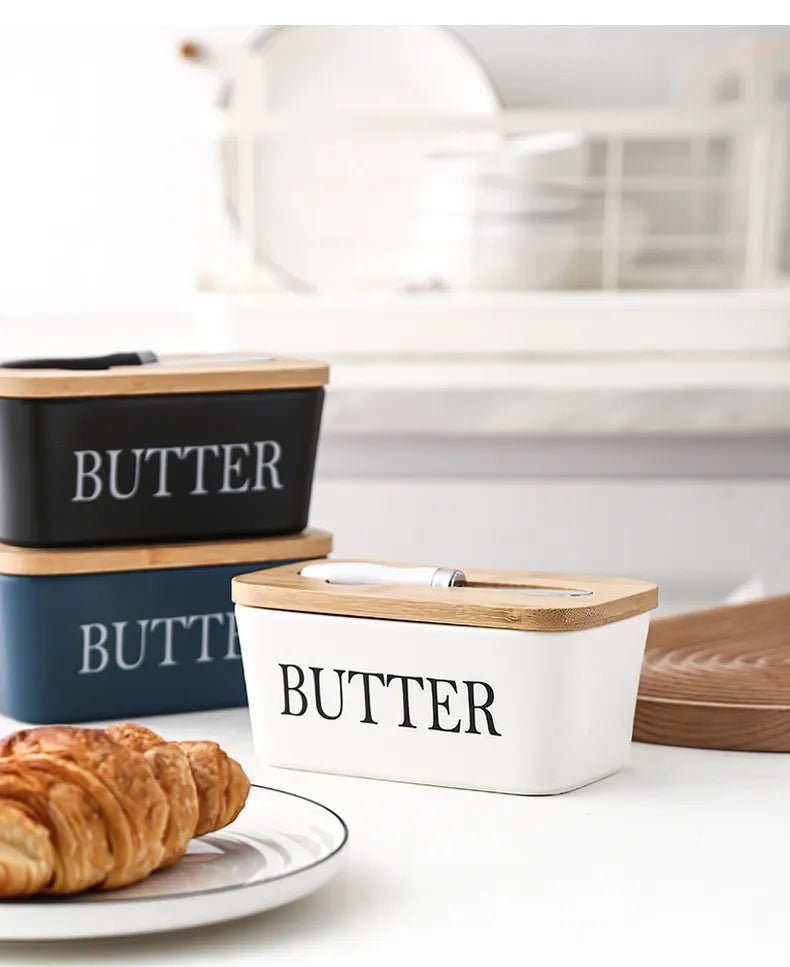 White ceramic butter dish filled with butter, shown with stacked black butter dishes and bamboo lids in modern kitchen