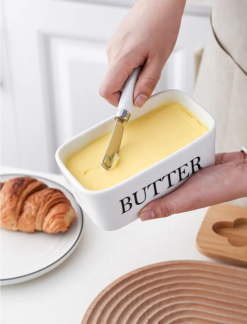 White ceramic butter dish with butter block beside a croissant on plate and stainless steel spreader, front view