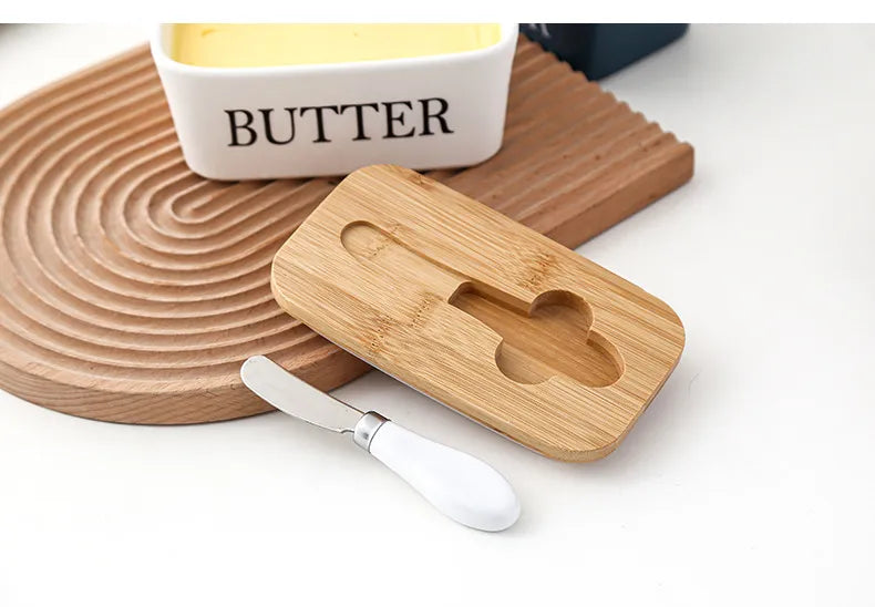White ceramic butter dish with butter block beside a croissant on plate and stainless steel spreader, side view